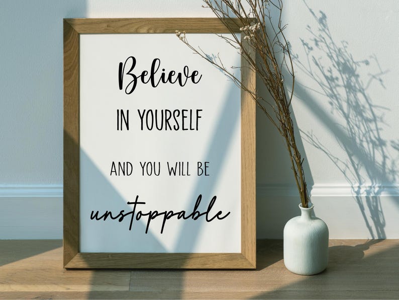 K&ouml;nnte beinhalten: Ein gerahmter Druck mit den Worten "Believe in Yourself and You Will Be Unstoppable" in schwarzer Schrift. Der Rahmen ist aus hellem Holz. Rechts vom Rahmen steht eine kleine wei&szlig;e Vase mit getrockneten Zweigen.