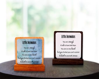 May include: Two desk signs with the text "Little Reminders" and inspirational quotes. One sign has a light wood base and a blue watercolor background. The other has a dark wood base and a white background.