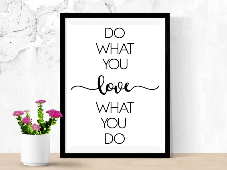 May include: A framed print with the text "DO WHAT YOU love WHAT YOU DO" in black against a white background. The frame is black. A small white vase with pink flowers sits in front of the print.