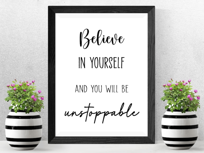 K&ouml;nnte beinhalten: Ein gerahmter Druck mit den Worten "Believe in Yourself and You Will Be Unstoppable" in schwarzer Schrift. Zwei dekorative Vasen mit schwarz-wei&szlig;en Streifen und kleinen rosa Blumen flankieren den gerahmten Druck.