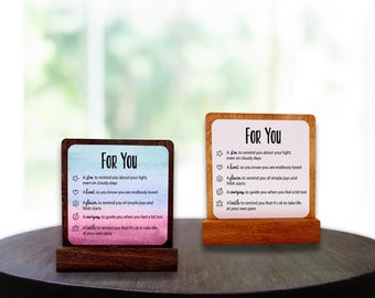 May include: Two wooden desk signs with the text "For You" and inspirational messages. The signs have a gradient background of blue and pink. The signs are on a dark wooden table.
