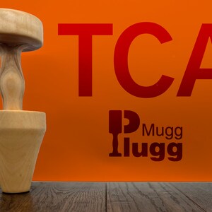 May include: A wooden tamper with a round top and a cone-shaped base. The tamper is sitting on a wooden surface. The text "TCA Mugg Ilugg" is visible in the background.