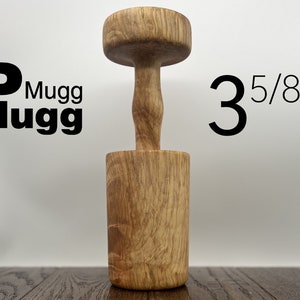 Peut inclure: Un bouchon de tasse en bois, d'environ 9,2 cm de haut, avec une base cylindrique et une poignée. Le produit est en bois clair et porte le texte "Mugg Plugg" en noir. L'article est conçu pour boucher une tasse.