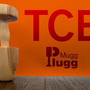 May include: A wooden mallet with a round head and a handle. The mallet is sitting on a wooden surface. The background is orange with the text "TCB" in red letters and the text "Mugglugg" in red letters.