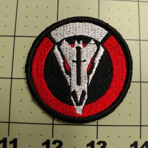 May include: A black and white embroidered patch with a skull design. The skull is surrounded by a red circle with a white outline. The patch is about 2 inches in diameter.