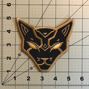 May include: Black and gold embroidered patch of a stylized cat head with glowing eyes.