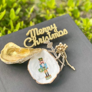 May include: A gold painted oyster shell with a nutcracker illustration inside. The shell is sitting on a black surface with a gold glitter "Merry Christmas" sign and a gold twig with gold berries.