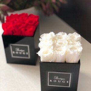 May include: Two black square boxes with lids, each containing a bouquet of preserved roses. The box on the left contains red roses and the box on the right contains white roses. Both boxes have a white label with the text "Boutique Rouge".