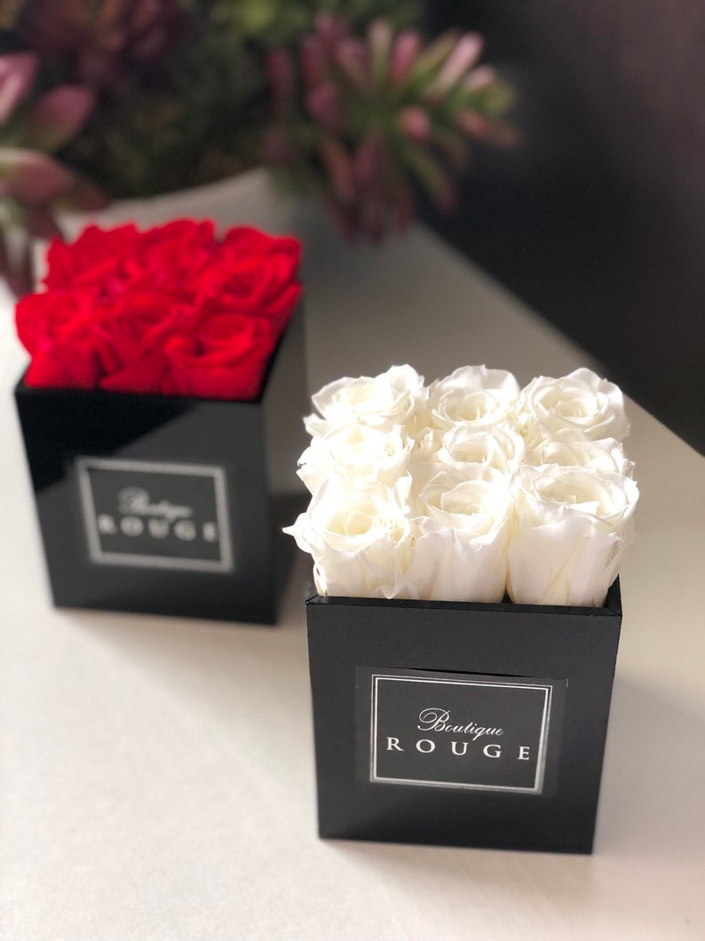 May include: Two black square boxes with lids, each containing a bouquet of preserved roses. The box on the left contains red roses and the box on the right contains white roses. Both boxes have a white label with the text "Boutique Rouge".
