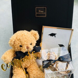 May include: A black gift box with a gold teddy bear holding a gold "Congrats" sign. The box also contains a framed picture with a graduation cap and a clear box with two gold roses.