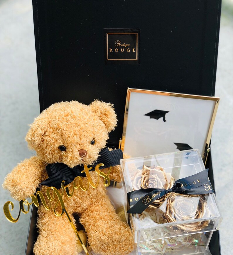 May include: A black gift box with a gold teddy bear holding a gold "Congrats" sign. The box also contains a framed picture with a graduation cap and a clear box with two gold roses.