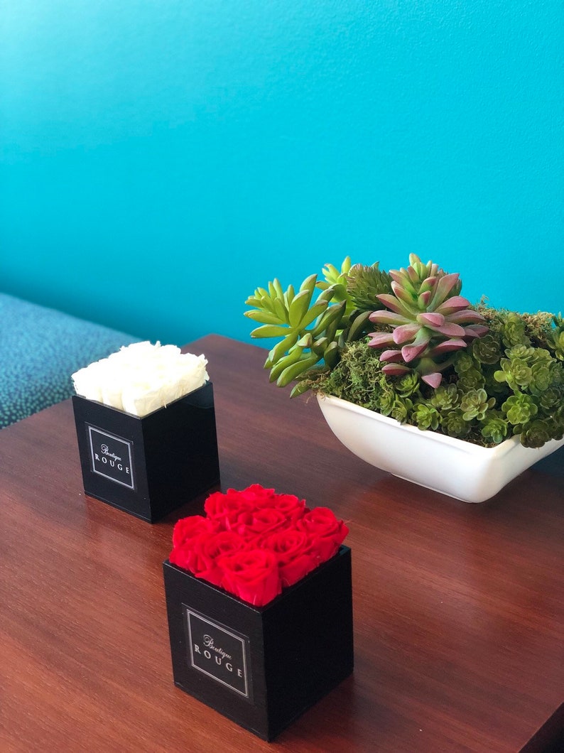 May include: Two black square boxes with white and red roses inside. The boxes have the text "Boutique Rouge" on the front. The boxes are on a wooden table with a white bowl of succulents in the background.