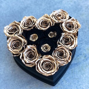 May include: A heart-shaped box filled with preserved roses. The roses are a mix of gold and black.