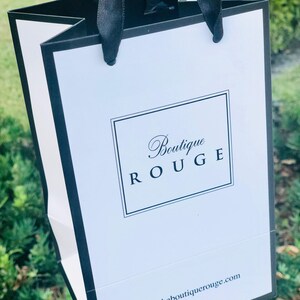 May include: A white shopping bag with black handles and trim. The bag has the text "Boutique Rouge" in a black script font and the website address "www.theboutiquerouge.com" printed on the front.