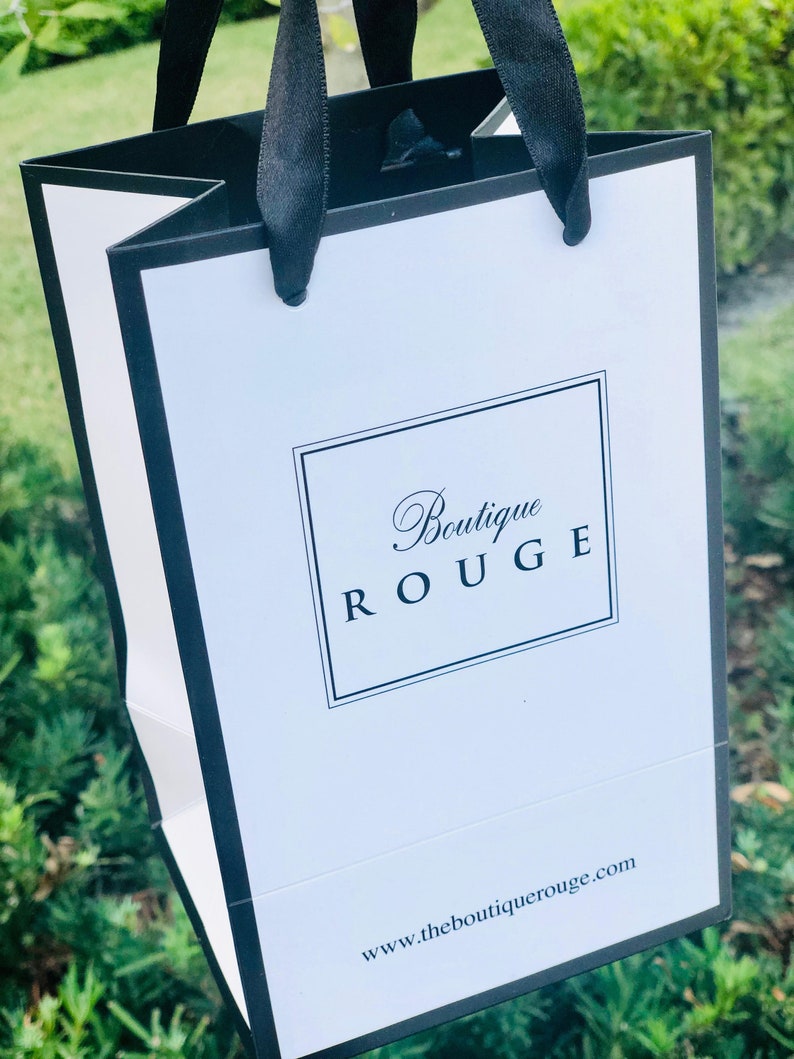 May include: A white shopping bag with black handles and trim. The bag has the text "Boutique Rouge" in a black script font and the website address "www.theboutiquerouge.com" printed on the front.