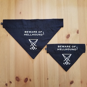Könnte beinhalten: Zwei schwarze Hunde-Bandanas mit weißem Text und einem Symbol. Die Bandanas sind dreieckig, mit den Worten "BEWARE OF HELLHOUND" oben aufgedruckt. Unter dem Text befindet sich ein weißes Symbol. Die Bandanas liegen auf einer Holzoberfläche.