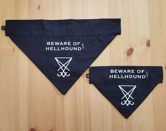 May include: Two black dog bandanas with white text and a symbol. The bandanas are triangular, with the words "BEWARE OF HELLHOUND" printed across the top. Below the text is a white symbol. The bandanas are on a wooden surface.