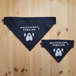 Könnte beinhalten: Zwei schwarze Hundehalstücher mit weißem Text und einer Grafik eines Pentagramms und Zähnen. Das größere Halstuch sagt "Malevolent Familiar" und das kleinere Halstuch sagt "Malevolent Familiar".