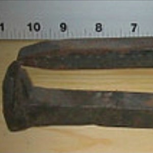 May include: Two rusty metal railway spikes, one is slightly longer than the other, are lying on a wooden surface next to a ruler.