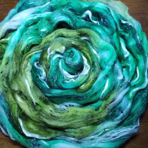 Può includere: Una disposizione circolare di fibra verde, turchese e bianca. La fibra è un mix di verdi chiari e scuri, con striature turchesi e bianche. La consistenza appare morbida e soffice, con una leggera lucentezza. Probabilmente per progetti di artigianato o tessili.