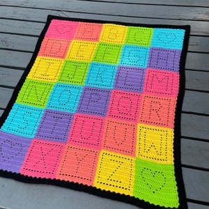 May include: A colorful crochet blanket with a black border. The blanket is made up of squares in various colors, including pink, yellow, green, blue, and purple. Each square has a heart design in the center.