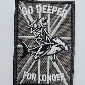 May include: Embroidered patch with a skeleton riding a hammerhead shark, holding a knife. The text "GO DEEPER FOR LONGER" is embroidered in white on a grey background. The patch has a black border.