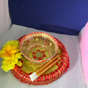 May include: A decorative serving set featuring a clear glass bowl and two chopsticks, resting on a gold and red embellished tray. A yellow flower is placed on the side. The set has a traditional, ornate design.