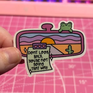 May include: A pink, blue, and yellow sticker with a cartoon sunset, a cactus, and a frog. The sticker is shaped like a rearview mirror and has a note attached that says "Don't look back, you're not going that way."
