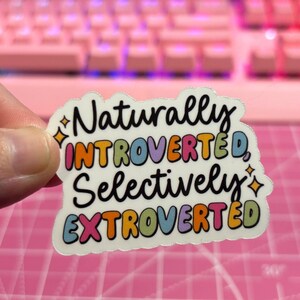 May include: A white sticker with a colorful, handwritten message that reads "Naturally Introverted, Selectively Extroverted". The sticker has a sparkly design and a wavy border.