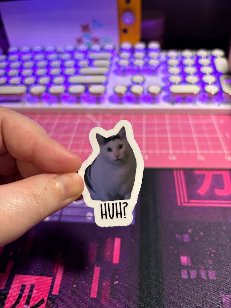 May include: A white and gray cat with black eyes looks at the camera with a confused expression. The cat is on a white sticker with the word "HUH?" printed in black below it.