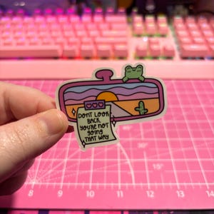 May include: A pink and purple car rearview mirror sticker with a sunset scene, a cactus, and a frog. The sticker has a note attached that says "DONT LOOK BACK. YOU'RE NOT GOING THAT WAY."