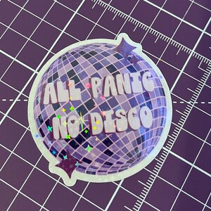 以下が含まれることがあります： 紫色のディスコボールのステッカーで、白い文字で「All Panic No Disco」と書いてあります。ステッカーはキラキラした仕上げで、紫色のグリッドの背景になっています。
