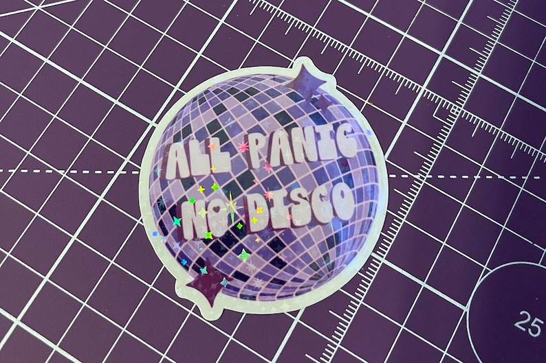 以下が含まれることがあります： 紫色のディスコボールのステッカーで、白い文字で「All Panic No Disco」と書いてあります。ステッカーはキラキラした仕上げで、紫色のグリッドの背景になっています。