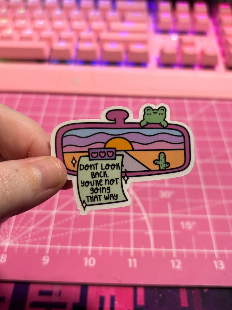 May include: A pink, blue, and yellow sticker with a cartoon sunset, a cactus, and a frog. The sticker is shaped like a rearview mirror and has a note attached that says "Don't look back, you're not going that way."