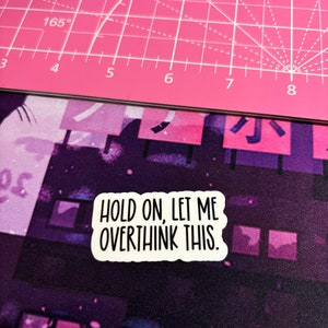 May include: A white sticker with the text "HOLD ON, LET ME OVERTHINK THIS." is placed on a purple and pink patterned surface. The sticker has a black border and is rectangular. The background includes a pink cutting mat.