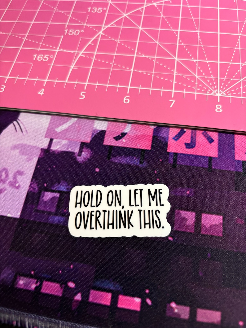 May include: A white sticker with the text "HOLD ON, LET ME OVERTHINK THIS." is placed on a purple and pink patterned surface. The sticker has a black border and is rectangular. The background includes a pink cutting mat.