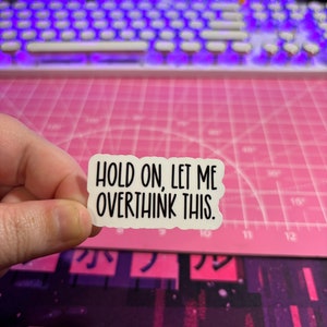 May include: A white sticker with black text that reads "HOLD ON, LET ME OVERTHINK THIS." The sticker is held by a hand against a pink cutting mat and a purple and white keyboard.