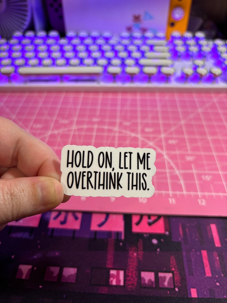 May include: A white sticker with black text that reads "HOLD ON, LET ME OVERTHINK THIS." The sticker is held by a hand against a pink cutting mat and a purple and white keyboard.
