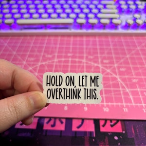 May include: A white sticker with the text "HOLD ON, LET ME OVERTHINK THIS." The sticker has a torn edge design. The background includes a pink cutting mat, a keyboard with purple lights, and a purple and black patterned surface.