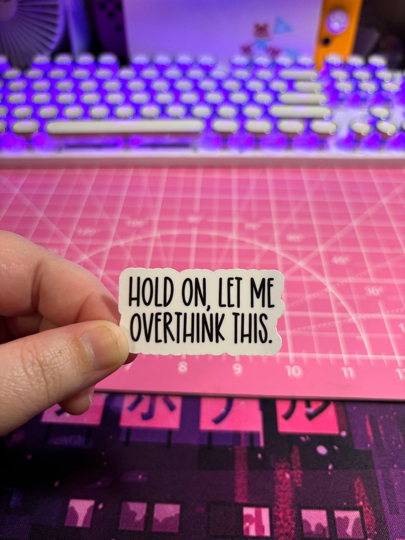 May include: A white sticker with the text "HOLD ON, LET ME OVERTHINK THIS." The sticker has a torn edge design. The background includes a pink cutting mat, a keyboard with purple lights, and a purple and black patterned surface.