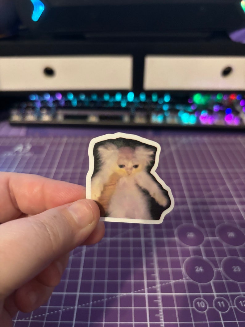 May include: A sticker featuring a digitally altered image of a cat with a surprised expression and outstretched arms. The cat has fluffy white fur and is set against a black background. The sticker is held by a hand against a purple cutting mat.