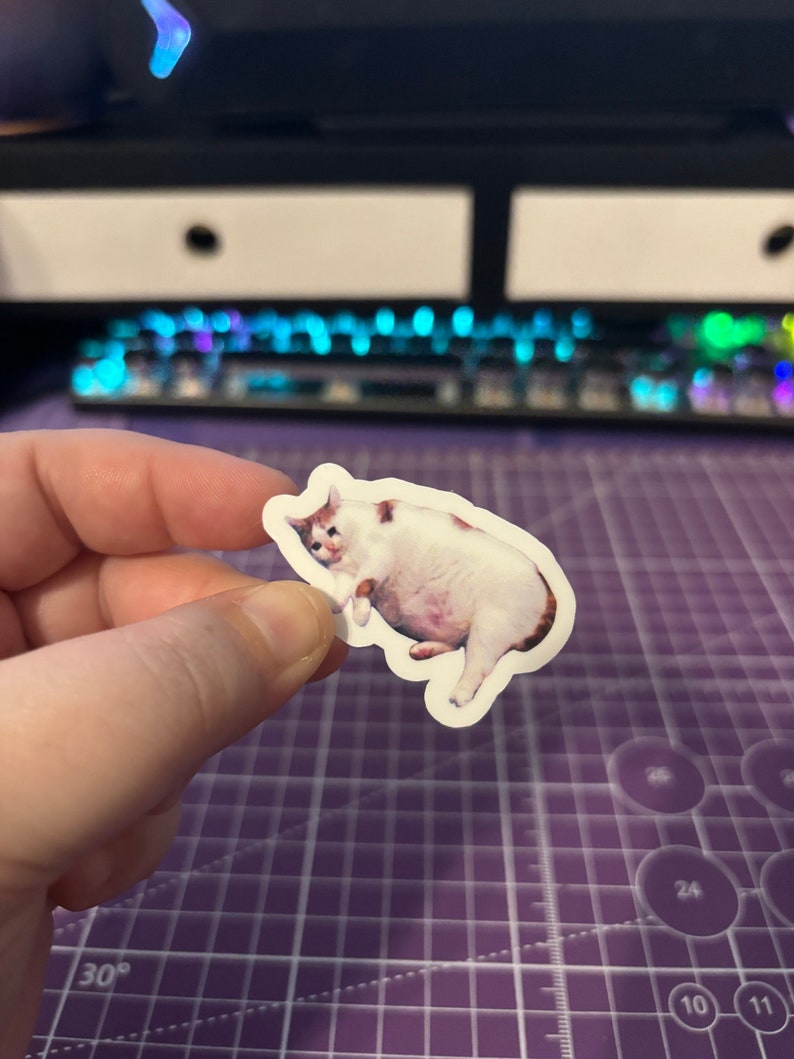 May include: A sticker featuring a meme of a sad-looking white cat with brown markings. The cat is depicted with a rounded body and a forlorn expression. The sticker is held against a purple cutting mat, with a keyboard in the background.