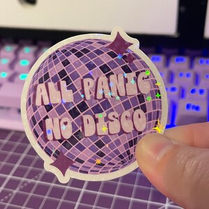以下が含まれることがあります： レトロなフォントで「All Panic No Disco」と書かれた、紫と白のディスコボールステッカー。ステッカーはキラキラした仕上げになっています。