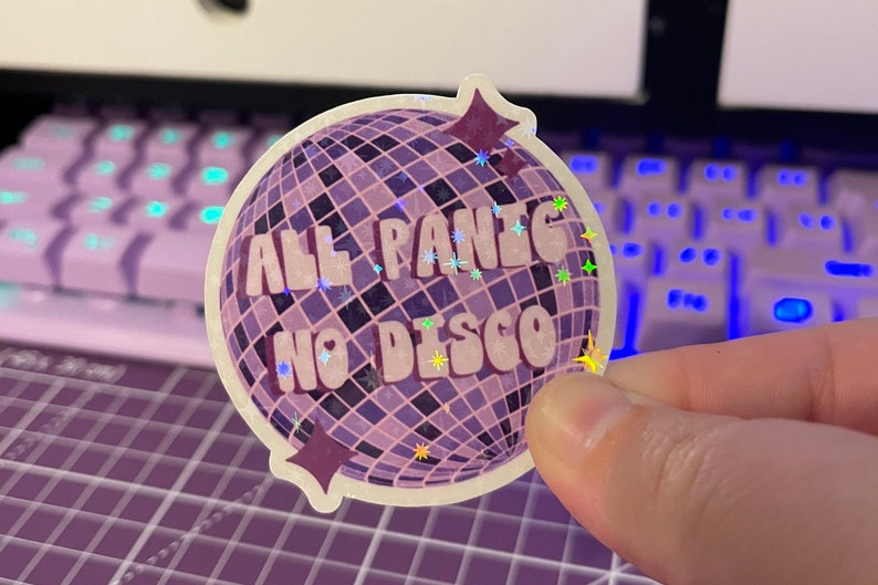 以下が含まれることがあります： レトロなフォントで「All Panic No Disco」と書かれた、紫と白のディスコボールステッカー。ステッカーはキラキラした仕上げになっています。
