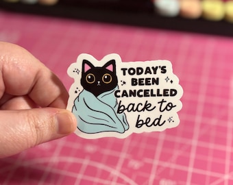 May include: A sticker featuring a black cat wrapped in a light blue blanket. The sticker has the text "TODAY'S BEEN CANCELLED back to bed" in a white font with star accents. The sticker is held by a hand.