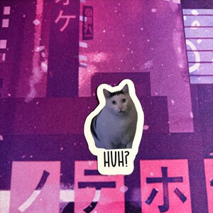 May include: A sticker of a cat with a white stripe on its face, looking confused. The sticker is on a purple background with Japanese characters and the text "HUH?" below the cat.