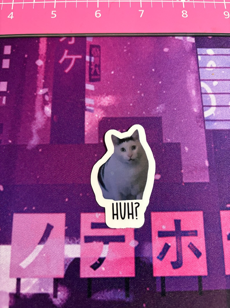 May include: A sticker of a cat with a white stripe on its face, looking confused. The sticker is on a purple background with Japanese characters and the text "HUH?" below the cat.