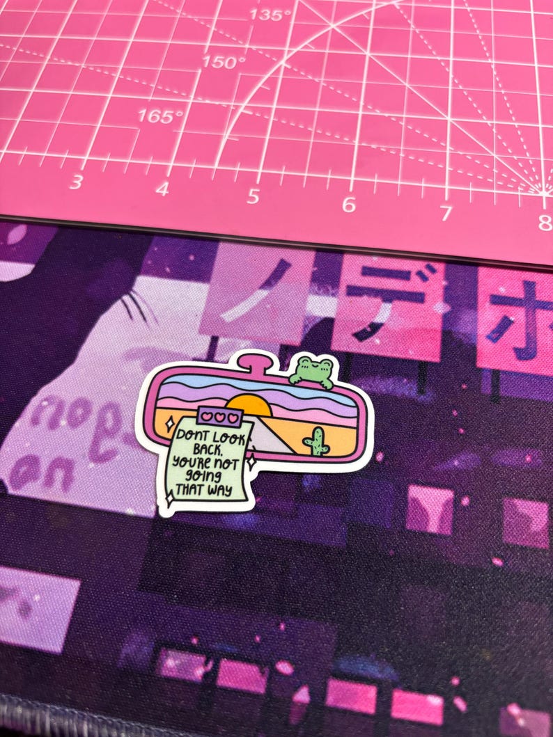 May include: Pink and green car rearview mirror sticker with a sunset and cactus scene. The sticker has a note that reads "Don't look back. You're not going that way."