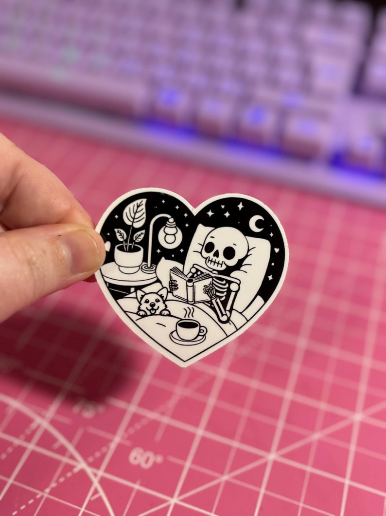 May include: A black and white sticker of a skeleton in bed reading a book with a small dog beside it. The sticker is in the shape of a heart and has a night sky background with stars and a crescent moon.