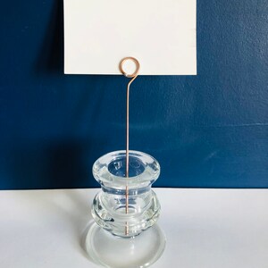 May include: A clear glass candle holder with a rose gold metal wire card holder. The card holder is attached to the top of the candle holder with a loop.